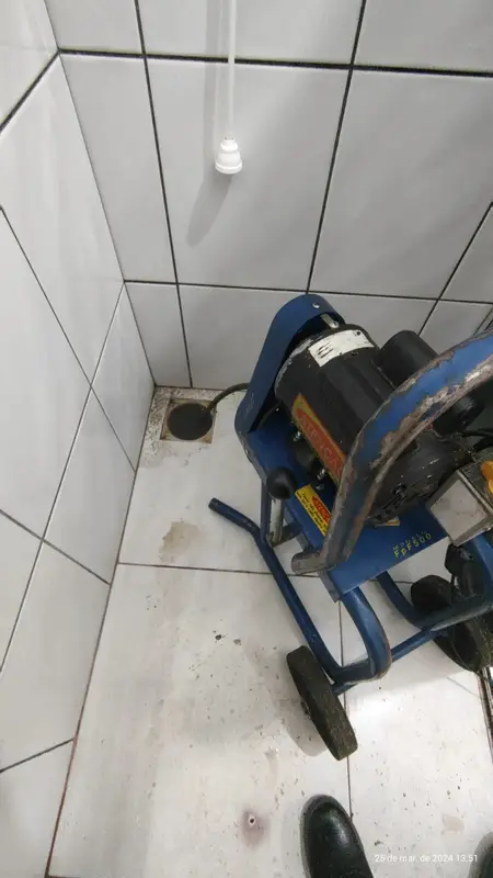 Máquina de desentupimento ao lado de ralo de banheiro com piso de azulejo branco